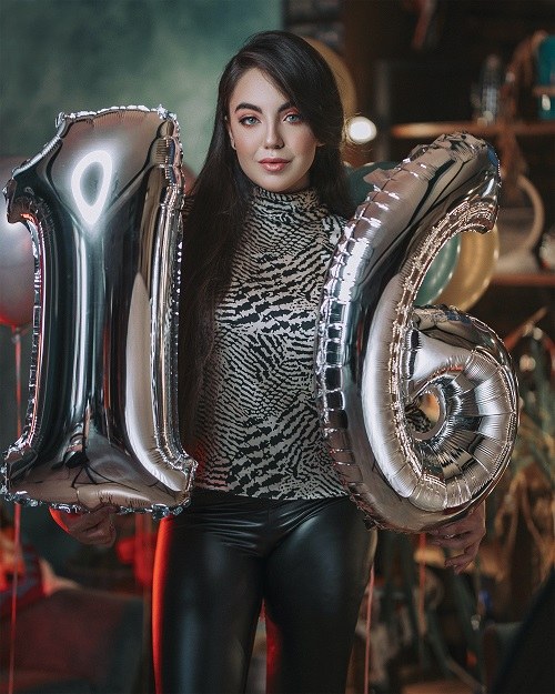 Dívka v tygřím tričku držící stříbrné balónky ve tvaru číslic jedna a šest. 