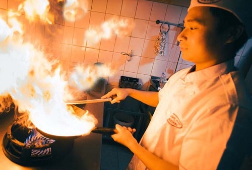 Asijský kuchař flambující jídlo na pánvi.