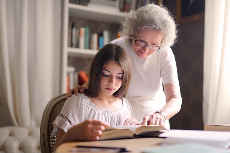 Vnučka čte v knize, babička se nad ní sklání a něco jí ukazuje.