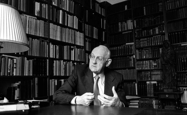 Černobílá fotografie Hanse Urse Von Balthasara sedícího v pracovně s knihami.