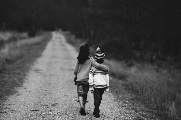 Černobílá fotografie dvou sourozenců, držících se za ramena a jdoucích po polní cestě.
