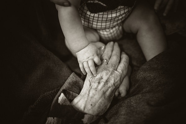 Černobílá fotografie staré dámské ruky, kterou drží malá ručka dítěte.