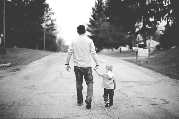 Černobílá fotografie s mužem otočeným zády držícím za ruku malého chlapce, s nímž jde po ulici. 
