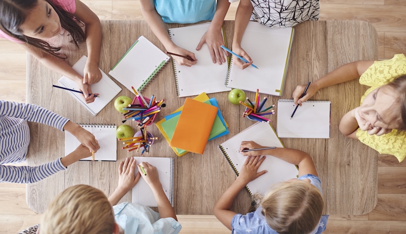 Děti si kreslí ve škole, na stole je mnoho pastelek a barevných sešitů.