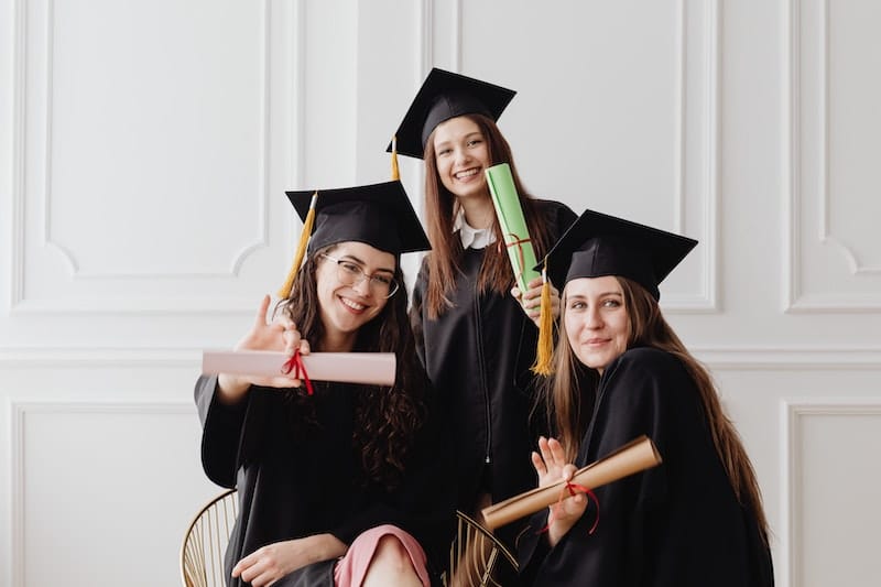 Tři dívky v záběru, po promoci, na sobě mají typické čepce, v rukou diplomy.