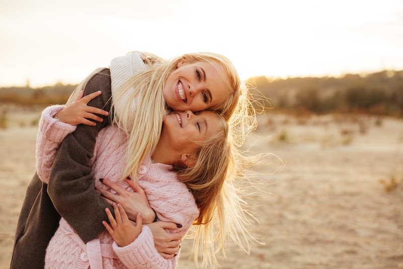 Matka a dcera na pláži, objímají se s úsměvem.