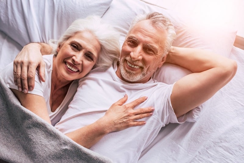 Starší manželský pár v bílém leží v posteli a usmívá se.