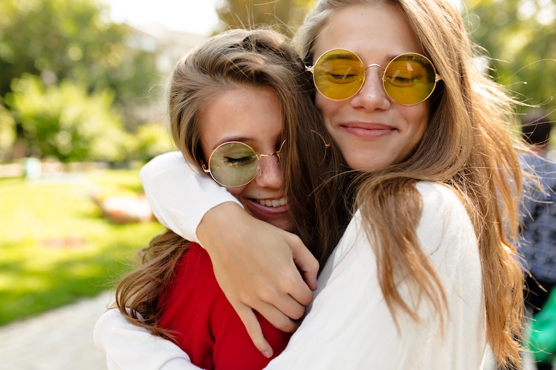 Dvě dívky ve slunečních brýlích se drží v pevném obětí, obě vypadají šťastně, usmívají se.
