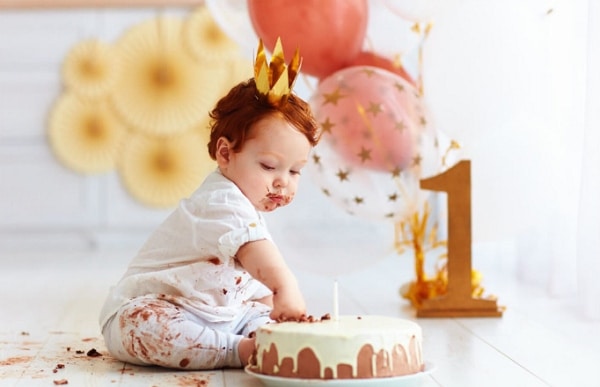 Čokoládou ušpiněné dítě, sedící na zemi se zlatou papírovou korunkou na hlavě, strkající ruku do narozeninového dortu. 