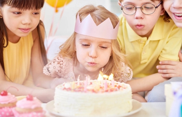 Dívka s korunkou z papíru na hlavě foukající na dort s narozeninovými svíčkami, obklopená skupinkou dětí. 