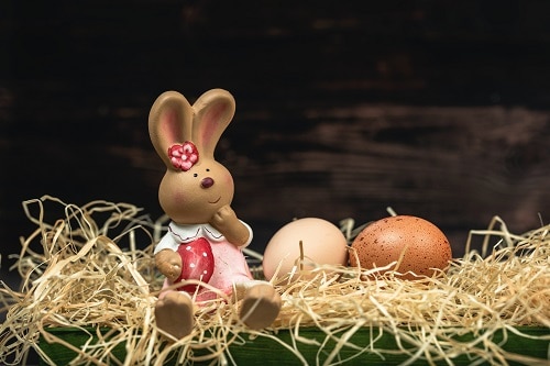 Keramická zaječí holčička, sedící ve slámě se dvěma hnědými vajíčky.