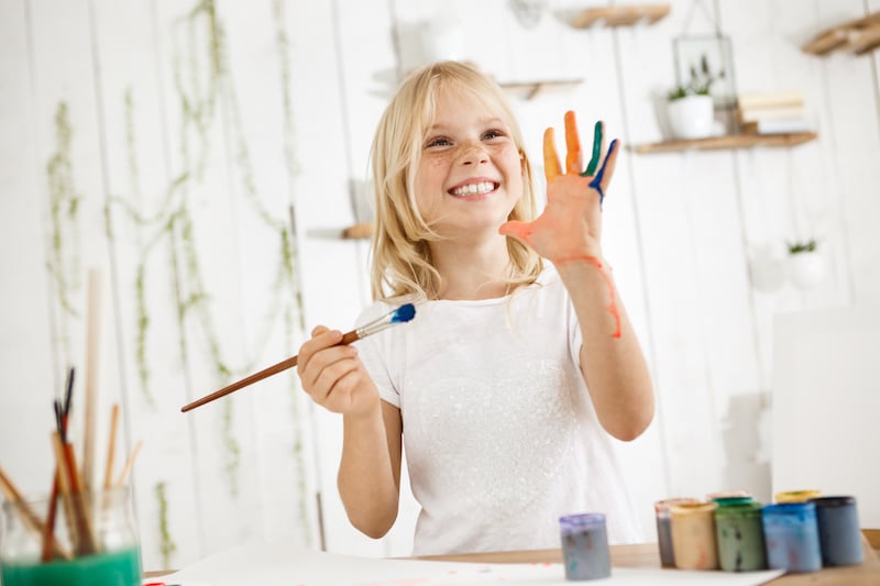 Blond holčička si maluje, drží štětec, druhou ruku má celou od barev a usmívá se.