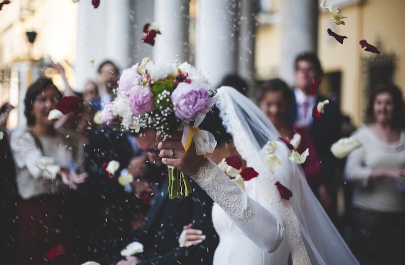 Svatebčané přímo po obřadu, hosté hází rýži, nevěsta drží kytici v čele obrazu.