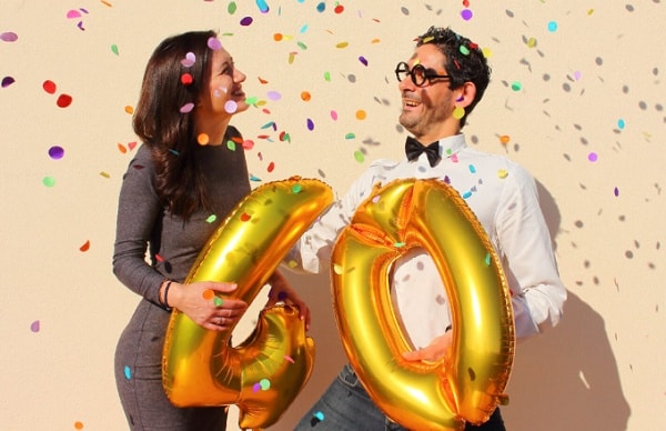 Smějící se muž se ženou, držící v rukou zlaté balónky ve tvaru čísla čtyřicet, obklopeni barevnými padajícími konfetami. 