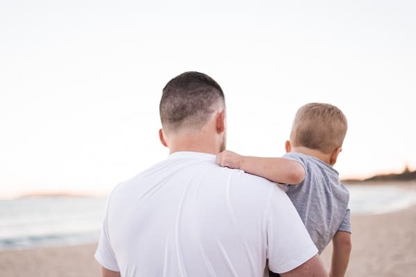 Muž otočený zády, držící v náručí malého chlapce na pláži.