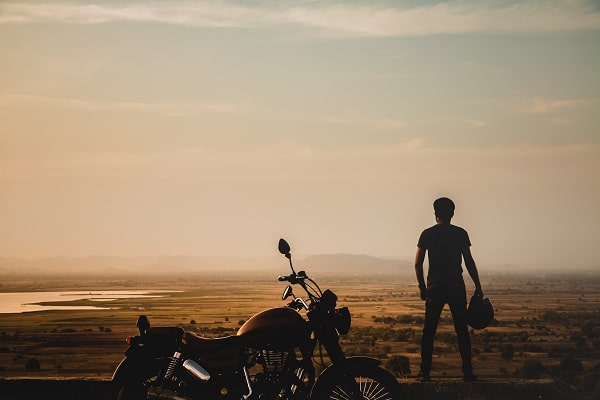 Muž stojící vedle motorky, sledující krajinu při západu slunce.