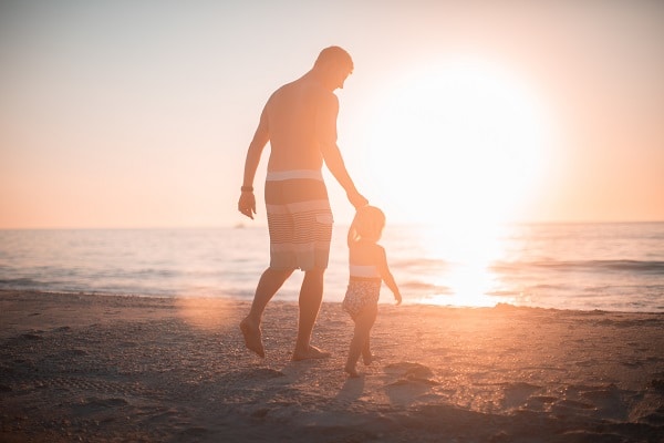 Otec vedoucí za ruku malé dítě po pláži u moře při západu slunce.