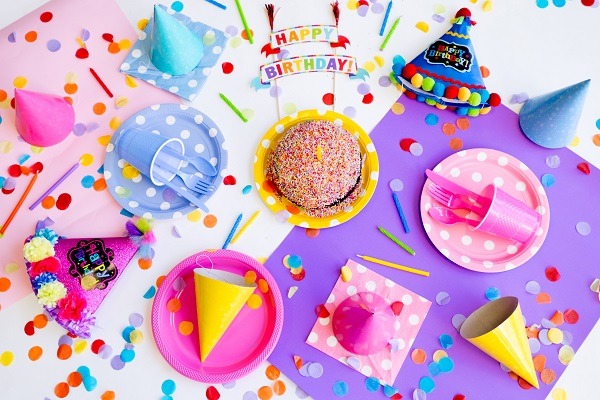 Barevná fotografie stolu s narozeninovým dortem, kelímky, narozeninovými čepičkami, svíčkami, konfetami a nápisem "Happy birthday". 