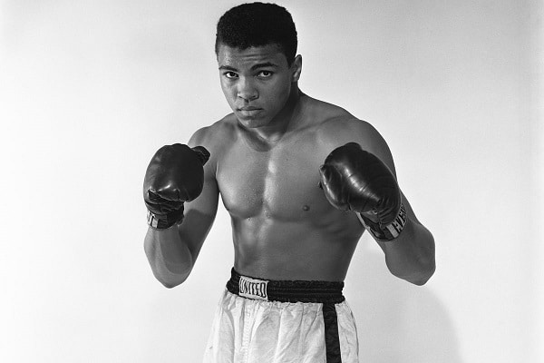 Černobílá fotografie Muhammada Aliho v bojovném postoji s boxerskými rukavicemi.