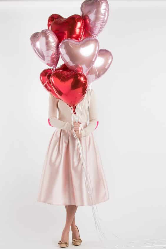 Žena v růžových šatech drží balónky ve tvaru srdíček. 