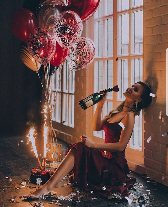 Oslavující holka v červených šatech s lahví šampaňského v ruce a s balonky kolem. 