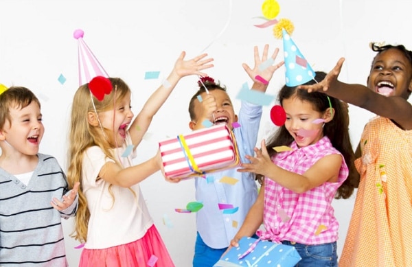 Jásající děti s narozeninovými čepičkami, dárečky, vyhazující konfety do vzduchu. 