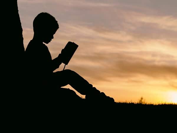 Silueta chlapce sedícího v přírodě čtoucího si knihu při západu slunce