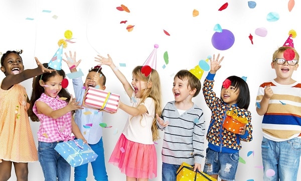 Skupinka dětí s narozeninovými čepičkami, dárečky, slavící narozeniny s létajícími konfetami.