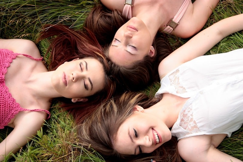 Tři ženy leží na trávě, mají zasněně zavřené oči, jedna se usmívá.
