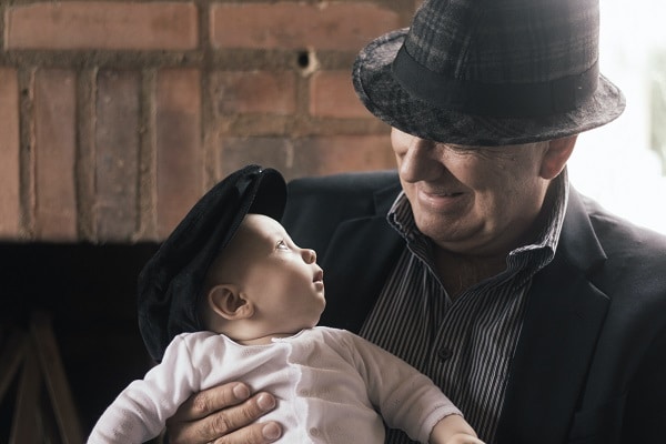 Starší smějící se muž s kloboukem držící v náručí malé, udiveně se koukající, miminko.