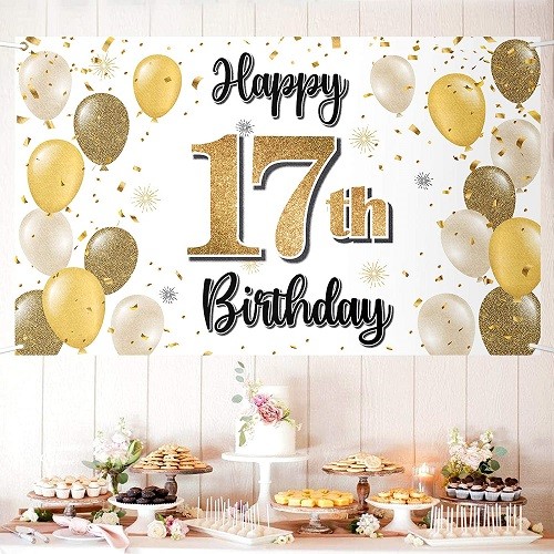 Stůl s dortem a dobrotami, nad nímž visí plakát se zlatým nápisem "Happy 17th birthday" a zlatými nafukovacími balónky. 