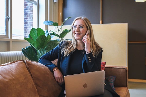 Smějící se žena sedící na gauči v kanceláři s mobilem u ucha a notebookem na klíně.