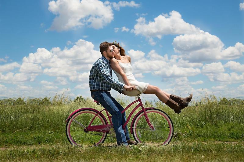 muž jede na růžovém kole, ženu veze na nosiči, krásný den, obloha s bílými mraky