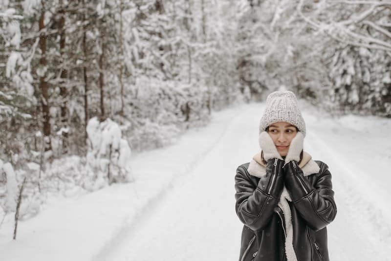 Dívka v bílé čepici v zasněžené krajině, rukavicemi si kryje tváře.